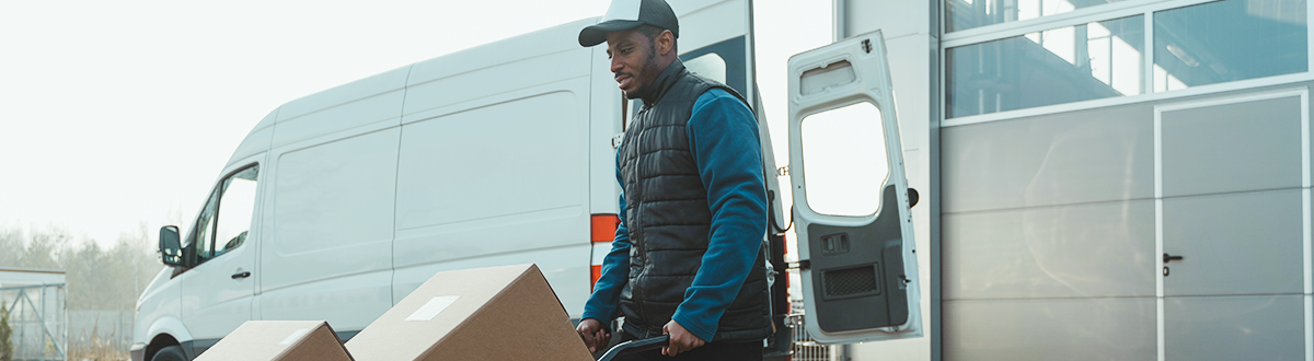 A man in a puffer vest moves boxes out of a commercial cargo van using a hand truck.