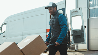 A man in a puffer vest moves boxes out of a commercial cargo van using a hand truck.