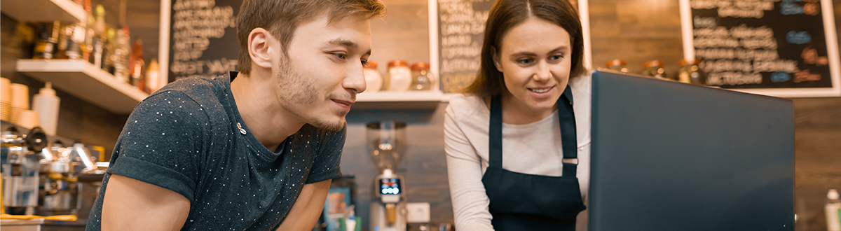 A young man and a young woman working at a coffee shop and looking at a laptop computer.