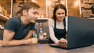 A young man and a young woman working at a coffee shop and looking at a laptop computer.