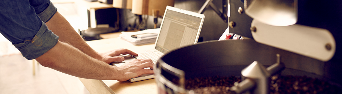 Man leaning on a counter using a laptop, surrounded by coffee and coffee roasting equipment.
