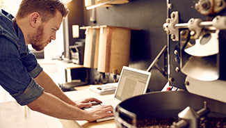 Man leaning on a counter using a laptop, surrounded by coffee and coffee roasting equipment.