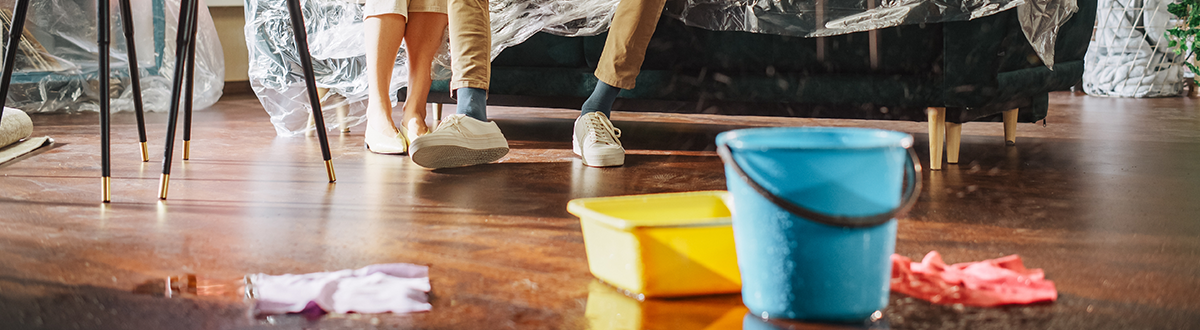 Close up of buckets filling with water from a leaking ceiling as water pools on a living room floor.