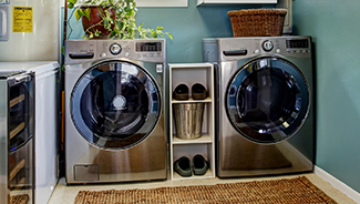 A brushed steel washer and dryer set in a home laundry room, with teal walls and a brown jute rug.