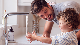 A man and a child smile as they wash their hands together under a running water tap in the kitchen.