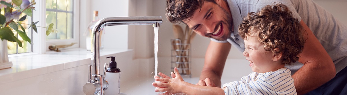 A man and a child smile as they wash their hands together under a running water tap in the kitchen.