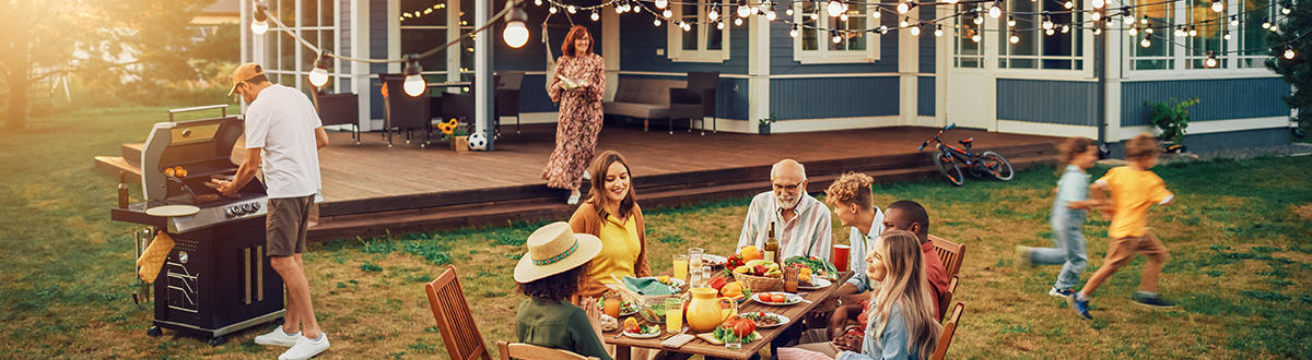 Family and friends are celebrating with an outdoor gathering in the yard of a large house. A man grills, a group sits at a table, and children play.