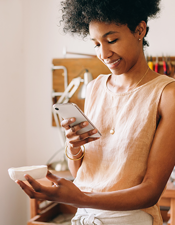 A woman in a tan sleeveless shirt smiles as she photographs a ceramic jewelry dish with her phone.