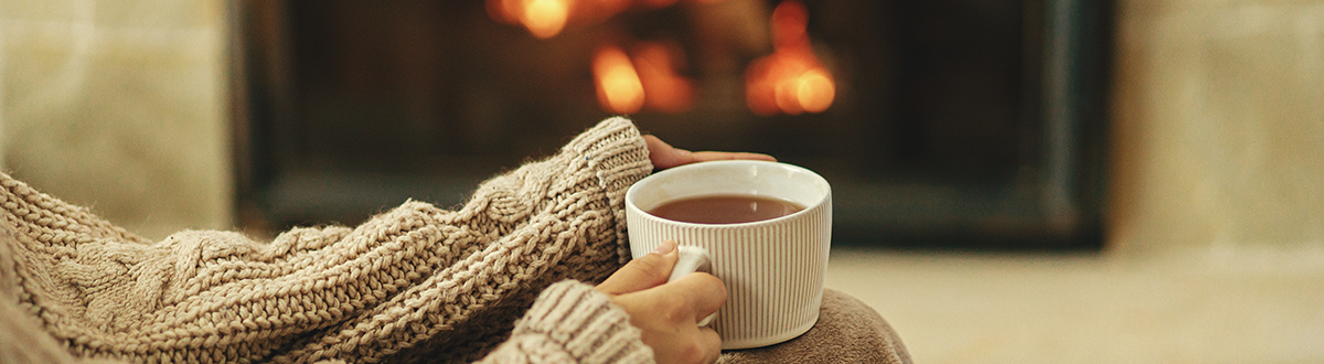 Hands in a cozy sweater hold a cup of warm tea with a burning fireplace in the background