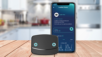 A LeakBot water leak detection device sits on a table next to a smart phone displaying the LeakBot app.